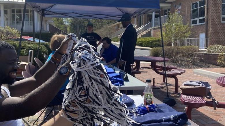 Arms wave pom poms while Chancellor Dr. A stands in the distance under a sun tent.